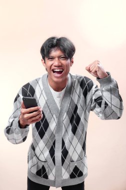 Asyalı adam akıllı telefonu heyecanla tutuyor, yumruğunu kaldırırken kendine güveniyor, mutluluk, motivasyon, başarı ve pozitif enerjiyi modern yaşam tarzı konseptinde gösteriyor..