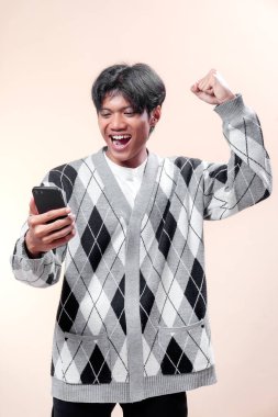 Akıllı telefonu tutarken heyecanla gülümseyen Asyalı adam, kutlama için yumruğunu kaldırıyor, mutluluk, güven, başarı, motivasyon ve pozitif enerjiyi ifade ediyor..