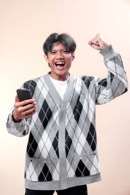 Asyalı adam tek eliyle akıllı telefonu tutarken heyecanla yumruğunu kaldırıyor, güvenle gülümsüyor, başarıyı, mutluluğu, motivasyonu ve pozitif enerjiyi ifade ediyor..