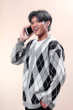 Asyalı adam akıllı telefonda konuşurken gülümsüyor, şık bir baklava desenli süveter giyiyor, kendinden emin ve pozitif bir ifade sergiliyor, modern iletişim kavramı..