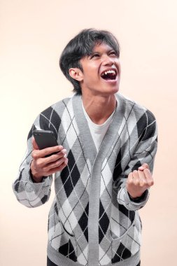 Asyalı adam akıllı telefonu neşeli bir ifadeyle tutuyor, başarıyı yumruğuyla kutluyor, baklava desenli süveter giyiyor, enerji ve mutluluk gösteriyor..