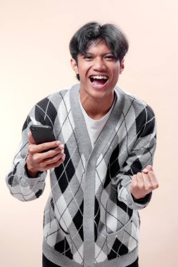 Asyalı adam elinde akıllı telefonuyla mutlu bir gülümsemeyle heyecan ve başarı sergiliyor. Modern moda tarzında şık bir süveter giyiyor..