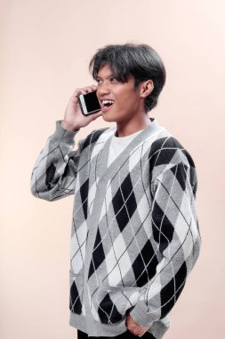 Akıllı telefonda konuşurken gülümseyen Asyalı bir adam, günlük baklava desenli süveter giyiyor, kendinden emin genç bir erkek pozitif ifade ve modern iletişim tarzı sergiliyor..