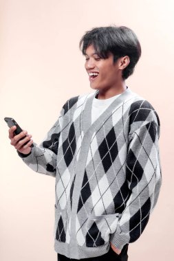 Asyalı adam akıllı telefona bakarken gülümsüyor, günlük baklava desenli süveter giyiyor, mutluluk ve olumlu duygu gösteriyor, modern yaşam tarzı ve dijital bağlantı..