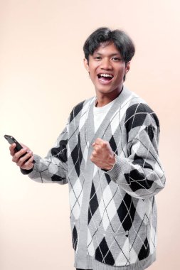 Asyalı adam akıllı telefonu neşeli bir ifadeyle ve sıkılmış yumrukla tutuyor, başarıyı, mutluluğu ve pozitif enerjiyi simgeliyor, iş, teknoloji, yaşam tarzı ve reklam kavramları için mükemmel..
