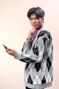 Asyalı adam gri baklava desenli süveter giyiyor. Akıllı telefonu ciddi bir ifadeyle tutuyor, çenesine dokunuyor düşünceli bir şekilde, kararı sembolize ediyor, odaklanıyor, iletişim kuruyor, modern yaşam tarzı ve profesyonel davranışları var..