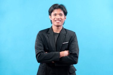 Siyah takım elbiseli kendine güvenen genç bir adam mavi bir arka plana karşı kollarını kavuşturarak gülümsüyor, iş kavramlarının profesyonelliğini, başarısını ve pozitif kişiliğini temsil ediyor..