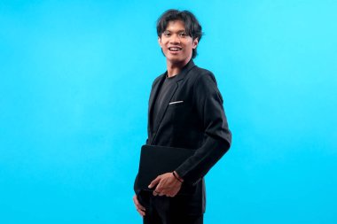 Siyah takım elbiseli, elinde dizüstü bilgisayarla gülümseyen genç adam, kendine güvenen ve şık görünüyor, mavi arka planda izole edilmiş, başarıyı, kariyer gelişimini ve pozitif iş davranışlarını temsil ediyor..