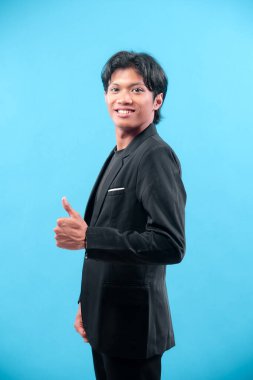 Siyah takım elbiseli kendine güvenen genç adam gülümsüyor ve mavi arka plana karşı olumlu bir hareket sergiliyor, başarıyı, pozitifliği ve profesyonelliği temsil ediyor..