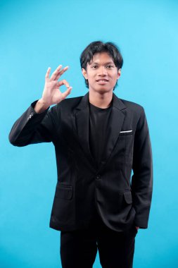Siyah takım elbiseli genç adam, mavi arka planda el hareketi yaparken güvenle gülümsüyor. Onayı, anlaşmayı, güveni, motivasyonu ve mesleki başarıyı simgeliyor..