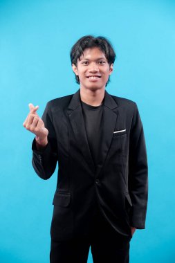 Siyah takım elbiseli genç adam mavi arka planda güven içinde gülümserken sevgi, ilgi, takdir, pozitiflik ve modern profesyonel tarzı sembolize ediyor..