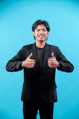 Siyah takım elbiseli gülümseyen genç adam mavi arka plana karşı çift parmak işareti gösteriyor, motivasyonu, cesareti, pozitifliği, güveni ve profesyonel başarıyı sembolize ediyor..