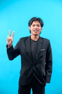 Siyah takım elbiseli genç adam mavi arka planda zafer işareti gösterirken özgüvenle gülümsüyor, başarıyı, pozitifliği, mutluluğu, kendine güveni ve profesyonel tutumu simgeliyor..