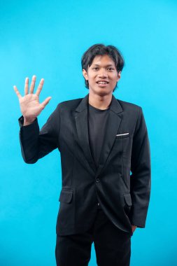 Siyah takım elbiseli genç bir Asyalı, dostça selamlaşarak olumlu iletişim ve profesyonel karizmayı ifade ederek elini kaldırırken kendine güvenerek gülümsüyor..