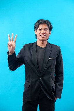 Siyah takım elbiseli kendinden emin genç adam gülümserken mavi arka planda üç parmak hareketi gösteriyor, üç numarayı sembolize ediyor, başarı, pozitiflik ve profesyonel tavır..
