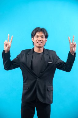Siyah takım elbiseli genç bir Asyalı, iki elini de barış işareti olarak kaldırarak güvenle gülümserken, pozitifliği, iletişimi ve profesyonel ifadeyi sembolize ediyor..