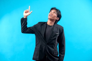 Siyah takım elbiseli ve tişörtlü genç bir adam işaret parmağıyla yukarı doğru bakarken, özgüvenini, merakını ve mesleki tutumunu mavi arka planda gösterir..