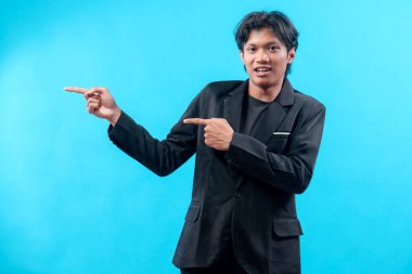 Siyah takım elbiseli ve tişörtlü genç bir adam iki eliyle kendine güveniyor, yön ve karizma gösteriyor, enerjiyi, pozitifliği ve mavi arka plandaki profesyonel tutumu temsil ediyor..