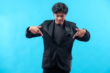 Siyah takım elbiseli genç bir adam iki eliyle aşağıya doğru gülümserken, özgüvenli bir jest, modern tarz ve parlak mavi bir arka plana karşı pozitif bir ifade sergiliyor..