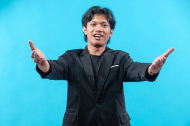 Siyah takım elbiseli genç bir adam iki elini de dostça bir gülümsemeyle öne uzatıyor. Mavi arka plana karşı misafirperverliği, iletişimi ve profesyonel tutumu sembolize ediyor..