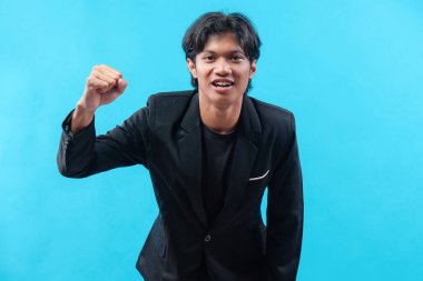 Siyah takım elbiseli genç bir adam, yumruğunu kaldırırken özgüvenle gülümser. Enerji, motivasyon ve azim gösterir. Olumlu ve profesyonel bir ifadeyle..