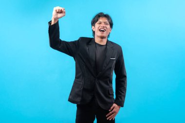 Siyah takım elbiseli neşeli genç bir adam yumruğunu kaldırırken kendine gülerek zaferi, hırsı, motivasyonu ve iş ve başarı kavramları için pozitif enerjiyi temsil ediyor..