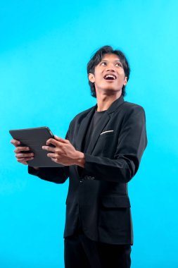 Siyah takım elbiseli genç bir Asyalı bir adam tablet tutuyor ve yukarı bakıyor. İlham verici bir ifadeyle, profesyonellik, yaratıcılık ve heyecanı modern bir iş bağlamında birleştiriyor..