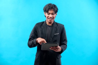 Siyah takım elbiseli gülümseyen Asyalı bir adam dijital tabletle tek parmakla, odaklanmış ve meşgul görünüyor, temiz mavi bir arka plana karşı güvenle duruyor..