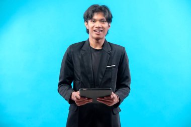 Siyah takım elbiseli, elinde tabletle kameraya gülümseyen, kendine güveni tam bir Asyalı genç adam, canlı mavi bir arkaplanda modern profesyonel bir görünüm sergiliyor..