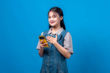 Sarı bir akıllı telefonu tutarken gülümseyen ve elini göğsüne koyan genç Asyalı kadın, minnettarlığını ya da mutluluğunu ifade ediyor, mavi bir arka plana karşı duruyor..