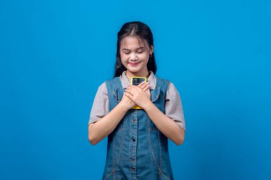 Gözleri kapalı genç bir Asyalı kadın tatlı tatlı gülümserken göğsüne sarı bir akıllı telefon sarılıyor, kot ve günlük kıyafetler giyiyor, stüdyoda koyu mavi bir arka planda izole edilmiş..