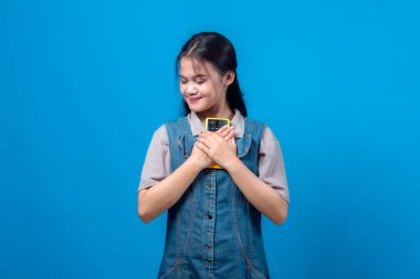 Gözleri kapalı genç bir Asyalı kadın tatlı tatlı gülümserken göğsüne sarı bir akıllı telefon sarılıyor, kot ve günlük kıyafetler giyiyor, stüdyoda koyu mavi bir arka planda izole edilmiş..