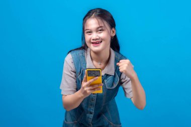 Neşeli, genç Asyalı bir kadın, elinde sarı bir akıllı telefon tutarken ve sevinçle gülümserken yumruk tokuştururken mavi bir arka plana karşı heyecan ve başarı sergiliyor..