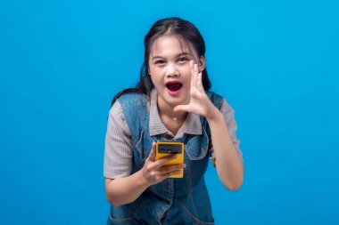 Heyecanlı genç Asyalı bir kadın, bir eli ağzının yanında bağırırken elinde sarı bir akıllı telefon tutarak heyecanını dile getiriyor ya da mavi bir arkaplan önünde bağırıyor..
