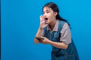 Genç Asyalı bir kadın, eli ağzının kenarında bağırıyor. Elinde bir akıllı telefon var. Heyecan ya da aciliyet gösteriyor..