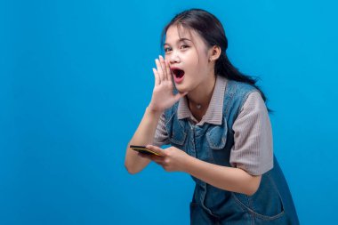 Neşeli, genç Asyalı bir kadın, eli ağızda bağırıyor, akıllı bir telefon tutuyor, heyecanını dile getiriyor ya da yüksek sesle bağırıyor, mavi stüdyo arka planına karşı duruyor..