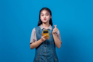 Sarı bir akıllı telefon tutan genç Asyalı kadın, bir parmak kalp hareketi yapıyor ve ördek yüz ifadesi, sağlam mavi bir arka plana karşı güvenle duruyor.