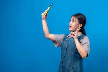 Heyecanlı genç Asyalı bir kadın, sarı akıllı telefonuyla selfie çekiyor, gülümsüyor ve eğlenceli bir el hareketi yapıyor, modern kıyafetin içinde parlak mavi bir arka plana karşı duruyor..