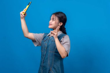 Genç Asyalı bir kadın, sarı akıllı telefonuyla selfie çekerken gülümsüyor. Parlak mavi bir arka plan önünde barış işaretiyle poz veriyor..