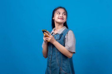 Genç Asyalı bir kadın iki eliyle sarı bir akıllı telefon tutarken gülümsüyor, umut dolu bir ifadeyle yukarı bakıyor, düz mavi arka planda sıradan bir kot elbise giyiyor..