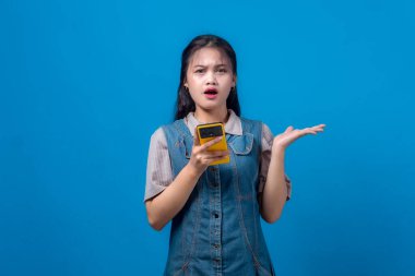 Genç Asyalı kadın şok olmuş ve kafası karışmış görünüyor sarı bir akıllı telefonu tutarken, bir elini inançsızlıkla kaldırırken. Beklenmedik haberlere duygusal tepki veriyor..