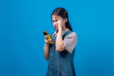 Üzgün, genç Asyalı bir kadın hayal kırıklığına uğramış görünüyor. Elinde sarı bir akıllı telefon, kaşlarını çatıyor ve tek eliyle gözünü ovuşturuyor. Mavi arka planda duruyor..