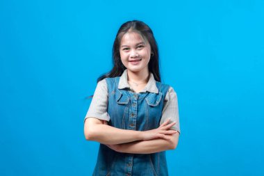 Uzun siyah saçlı, kendine güvenen, gülümseyen ve kollarını kavuşturan, mavi arka planda duran, kendine güvenen, pozitiflik ve iyimserlik gösteren kendine güvenen genç Asyalı bir kadın..