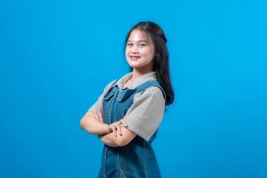 Uzun siyah saçlı, kendine güvenen, gülümseyen ve kollarını kavuşturan, mavi arka planda duran, kendine güvenen, pozitiflik ve iyimserlik gösteren kendine güvenen genç Asyalı bir kadın..