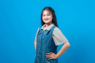 Mutlu, genç Asyalı bir kadın, elleri kalçasında, kot elbise giymiş ve sağlam mavi arka planda durarak güvenle gülümsüyor. Pozitif ifade, gündelik moda, stüdyo portresi.