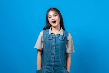 Saf mavi bir arka plan önünde poz veren Asyalı genç bir kadın. Sıradan bir kot pantolon ve kısa kollu gömlek giyiyor. Etkileyici, neşeli ve oyuncu görünüyor..
