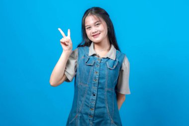 Genç Asyalı kadın gülümsüyor ve açık mavi bir arkaplanda dururken parmaklarıyla barış işareti gösteriyor. Yaşam tarzı veya gençlik temaları için neşeli ve olumlu ifade.