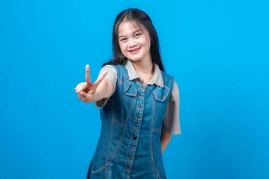 Genç Asyalı kadın gülümsüyor ve parmaklarını kameraya doğru uzatarak barış işareti yapıyor. Dost canlısı, kendine güveni olan, mavi arka planda günlük kot elbiseli, stüdyo portresi.