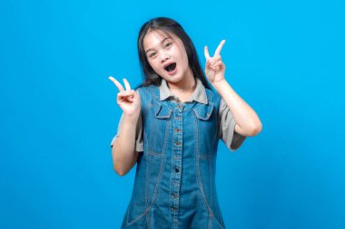 Ağzı açık gülümseyen ve iki elinde de barış işareti olan oyuncu, genç Asyalı kadın. İfade veren neşeli yüz, günlük kotlar, parlak mavi arka plan, stüdyo çekimi..
