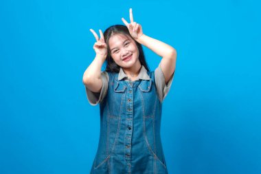 Neşeli genç Asyalı kadın gülümsüyor ve iki eliyle kafasının üstünde neşeli barış işaretleri yapıyor. Günlük kotlar, parlak mavi arka plan, neşeli yüz ifadesi, stüdyo çekimi..